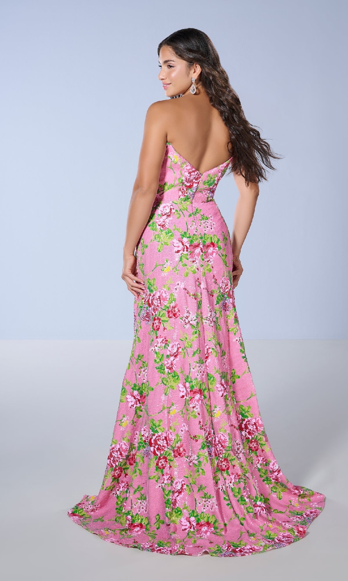 A woman wears the Tiffany 16143 long prom dress with a fitted waist, featuring a strapless floral design, as she stands and looks over her shoulder against a plain background.